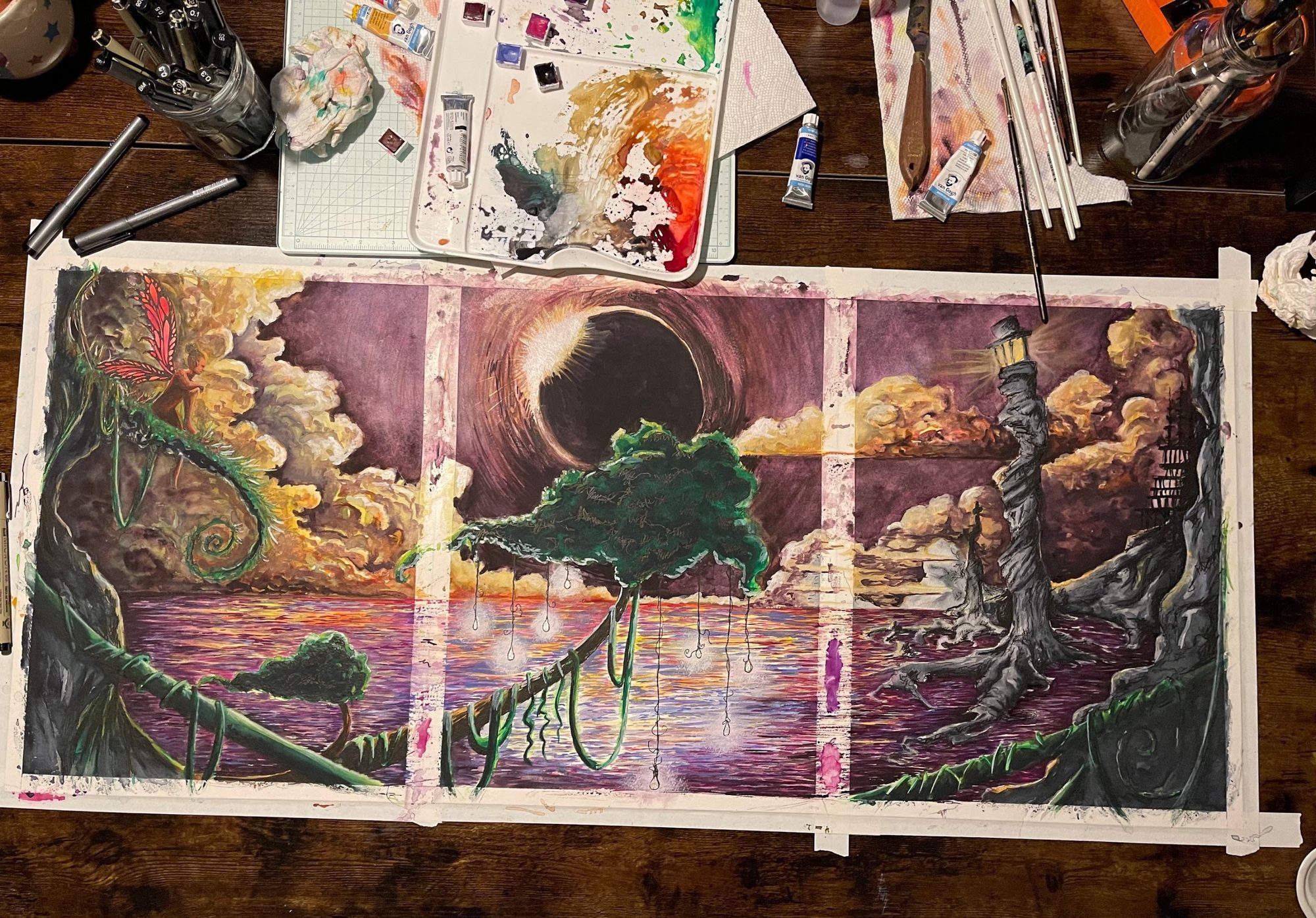 Overhead view of Justin's desk: a watercolor triptych of three 11 by 14 sheets taped side by side in the middle, surrounded by a paint-splattered palette, Van Gogh watercolor tubes, fineliner pens, brushes and paper towels