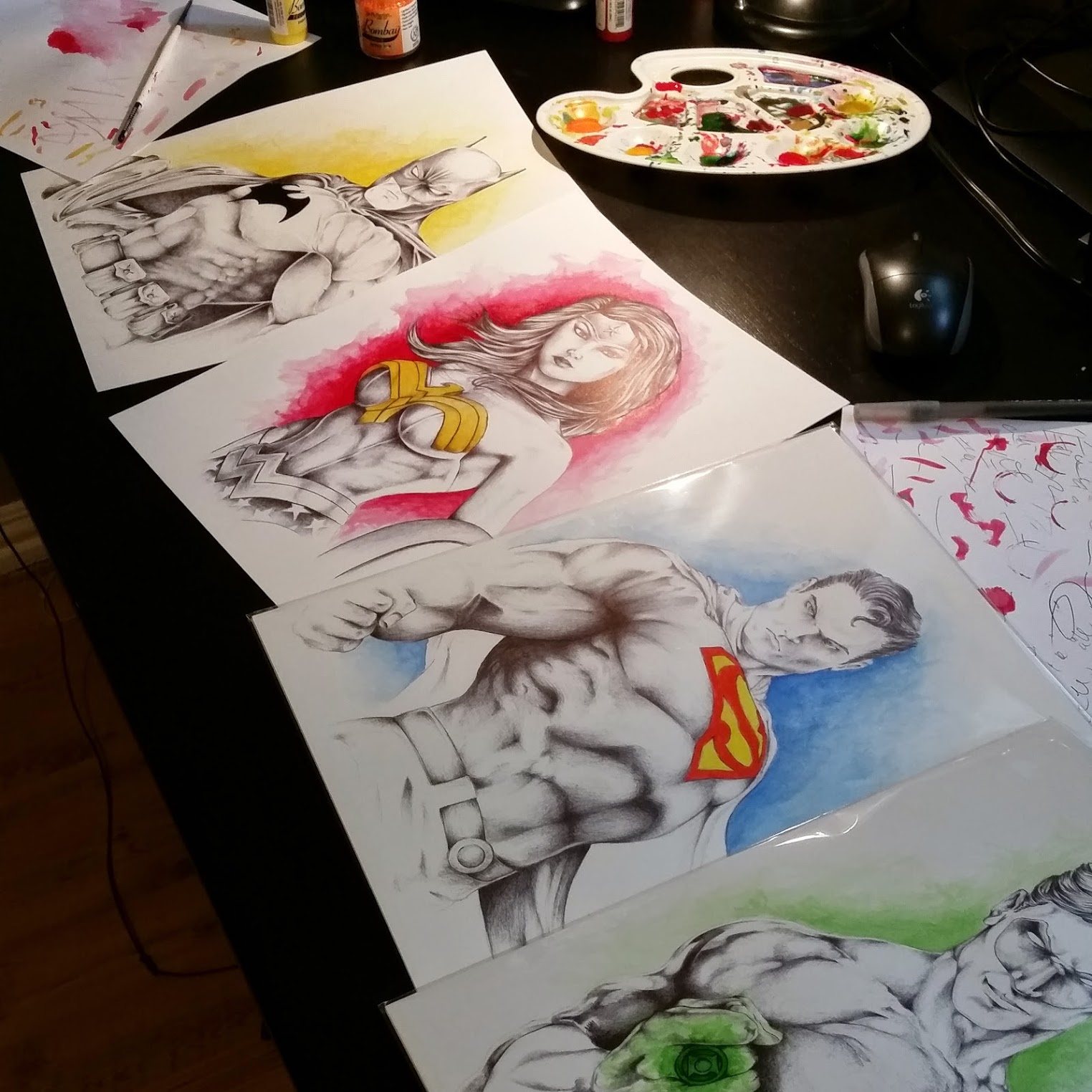 Batman, Wonder Woman, Superman and Green Lantern drawn in Bic pen with ink-wash backgrounds, laid out on a desk beside a paint palette