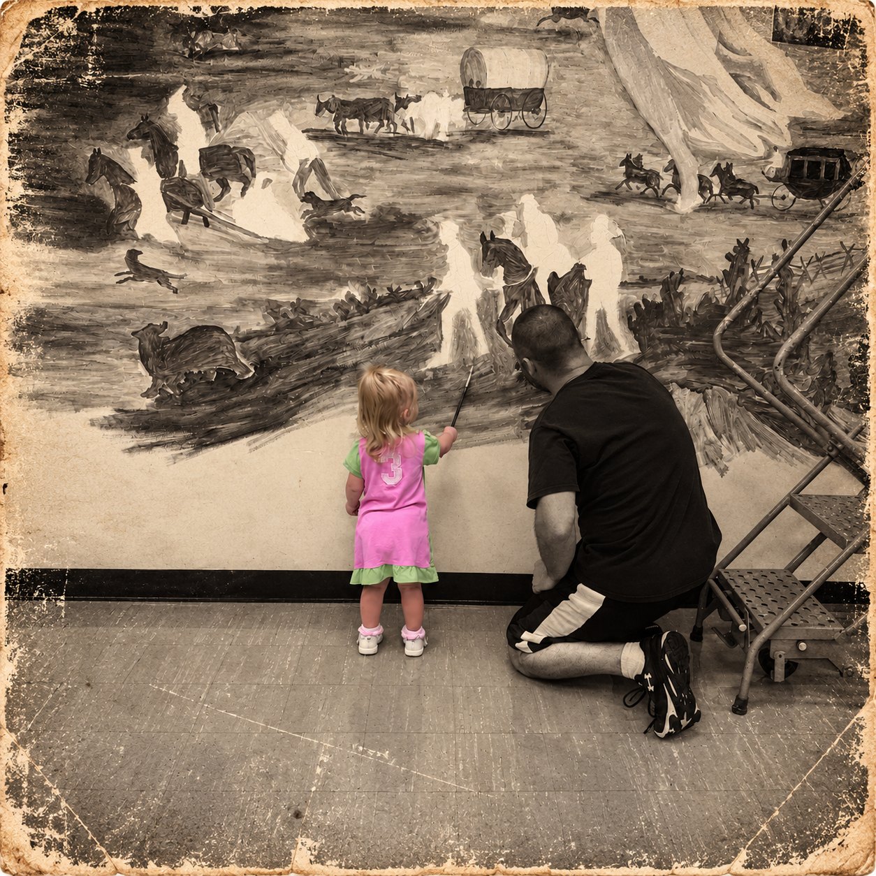 Justin kneels beside his young daughter, who holds a paintbrush up to a classroom mural of covered wagons, horses and a stagecoach crossing the plains